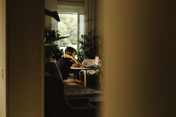 Teen sitting at table with laptop, studying or working at home, seen through doorway indoors
