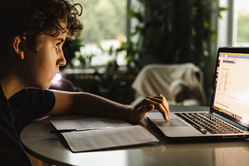 Teen boy studying at laptop, focused on screen with notebook and pen on table, evening light