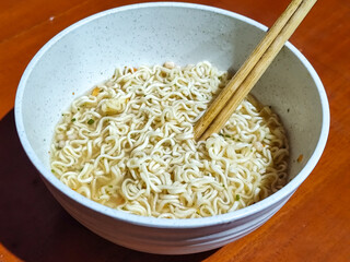 Chewy white instant noodles are held up in a white bowl, highlighting their texture and deliciousness, against the blurry background of the bowl.