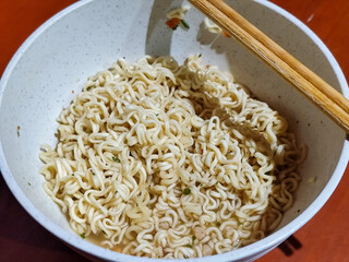 Chewy white instant noodles are held up in a white bowl, highlighting their texture and deliciousness, against the blurry background of the bowl.