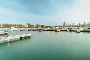 Photograph of a serene marina with numerous docks, calm water reflecting a clear sky, surrounded by bare trees, with a city skyline visible in the background.