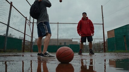 Two young men play basketball on a wet outdoor court with puddles and a cloudy sky in the background