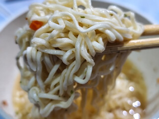 Rolls of springy white instant noodles are lifted with chopsticks from the bowl, highlighting their...