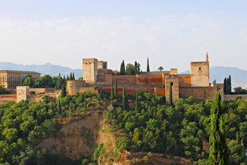 Fototapeta premium Panoramic view of the arabic fortress Alhambra in Granada from the Mirador de San Nicolás, showcasing the Nasrid palaces, towers, and ancient defensive walls rising above the lush forested hillside.