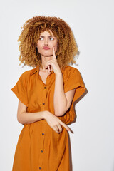 Young woman with curly hair in a thinking pose, wearing an orange button up dress against a white...
