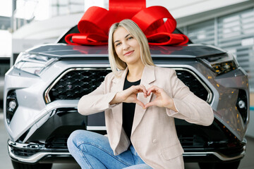 Woman making heart shape with hands in front of new car with red bow