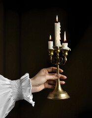 Human hand in white vintage shirts sleeve holds old brass candlestick with burning candles in the dark, vertical image.