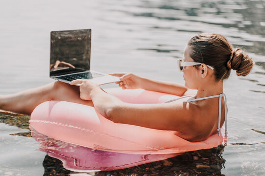 A woman is sitting in a pink inflatable raft on a lake, using a laptop. Concept of relaxation and leisure, as the woman is enjoying her time outdoors while working on her laptop.