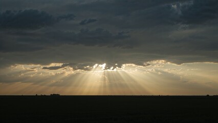 Golden sun rays pierce through the dramatic clouds over a dark field, creating a stunning and peaceful landscape at sunset