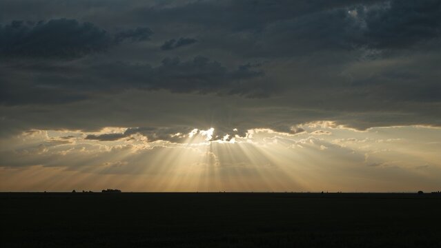 Golden sun rays pierce through the dramatic clouds over a dark field, creating a stunning and peaceful landscape at sunset
