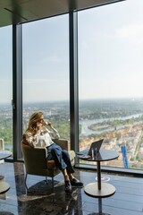 A businesswoman relaxes in a contemporary office setting, enjoying expansive views of the city skyline and river through large glass windows