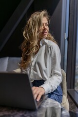 A fashionable woman with beautiful wavy hair is sitting at a sleek desk, deeply focused on her laptop in a modern workspace that boasts a stunning view of the city skyline in daylight