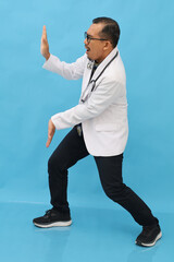 Full body of cheerful Asian adult male doctor wearing doctor's coat, stethoscope on neck, gesture pushing something aside in empty space, giving information, news or advertisement, blue background.