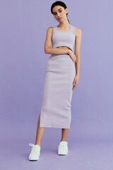 Young woman wearing casual lavender ribbed dress and white sneakers standing against pastel purple...