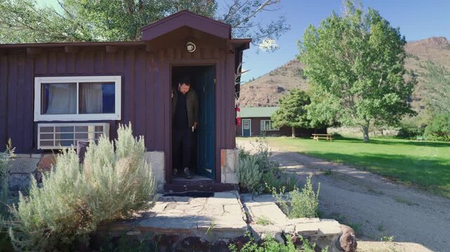 A man steps out of a cozy cabin nestled in a picturesque landscape, surrounded by serene nature, where rustic architecture beautifully complements the enchanting scenery of the countryside