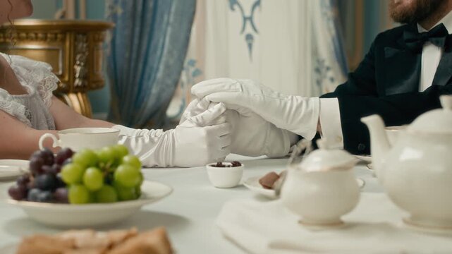 Cropped shot of aristocratic couple sharing tender moment, holding hands gracefully over refined tea setting, surrounded by fine china, fruit, and luxury decor