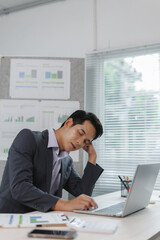 Tired office worker in a suit resting at a desk, struggling to stay awake, surrounded by charts and...