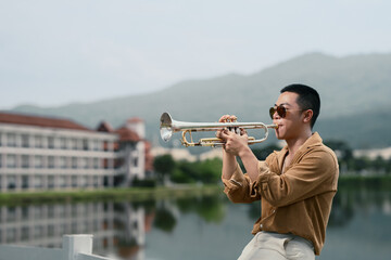Confident musician playing trumpet with serene water and mountain views.
