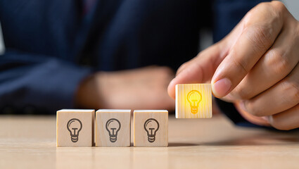 Wooden blocks with light bulb icons and a hand holding a glowing light bulb block on a table top
