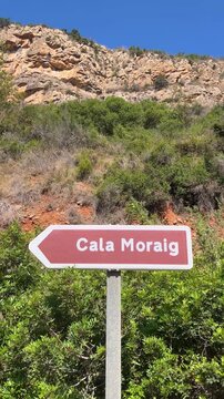 Brown tourist sign indicating the direction to Cala Moraig beach with cliffs and vegetation in the background, located in Benitachell, Alicante, Spain