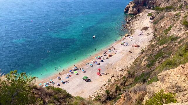 Tourists are swimming and sunbathing at Cala Moraig beach, a beautiful cove with crystal-clear turquoise waters located in Benitachell, Alicante, Spain, during a sunny summer day