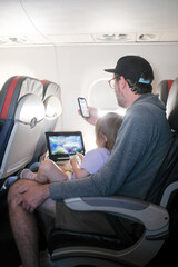 Toddler Daughter sits with Dad on Plane Watching Movie on a Tablet