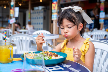 A young Japanese girl in a yellow dress eating rice and meat with a spoon at an outdoor beach café...