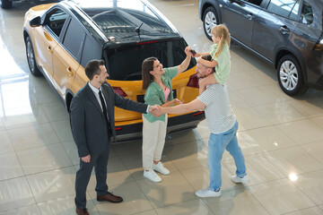 Salesman shaking hands with happy family near new car in salon