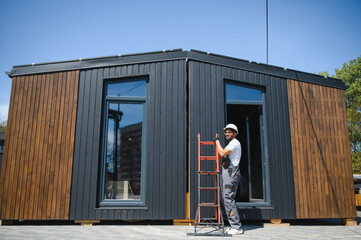 Construction worker placing ladder against modern prefabricated modular house
