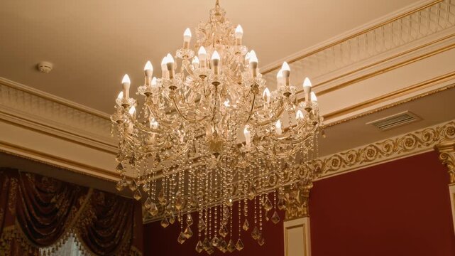 Grand crystal chandelier glowing warmly in richly decorated room with red walls, gold trim, and ornate ceiling and curtain details, radiating classic luxury and elegance