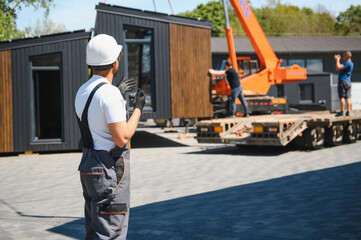 Construction worker supervising modular house transportation with crane and truck