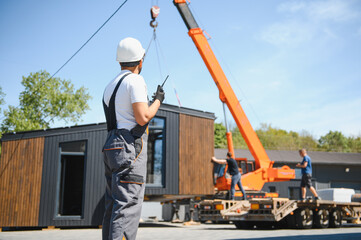 Construction worker supervising loading of modular house onto truck with crane