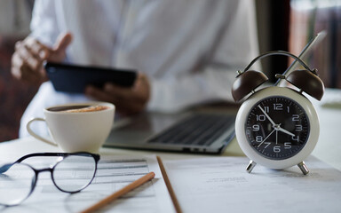 selective focus on alarm clock, asian businessman using laptop and tablet working on table before work time end in a day,  time management concept, routine work, office hour, blur background