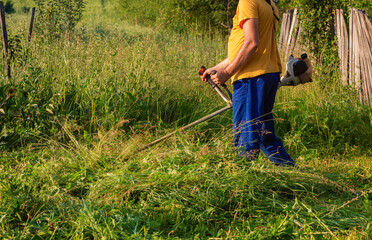 A man is trimming tall grass with a gas-powered weed trimmer near an wooden fence in backyard,...
