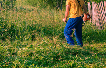 A man is trimming tall grass with a gas-powered weed trimmer near an wooden fence in backyard,...