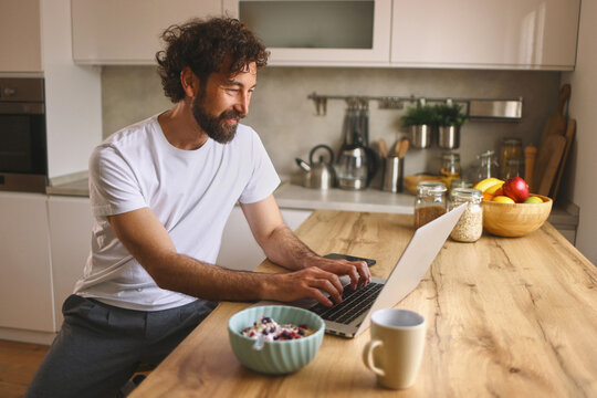 A man is sitting at a wooden kitchen table, typing on his laptop while having breakfast. He has a bowl of cereal and a cup of coffee beside him, showing a relaxed morning ambiance.