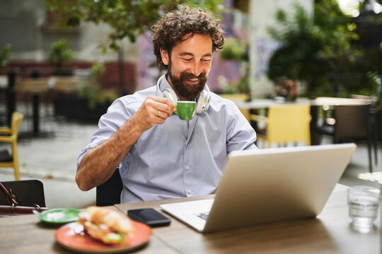 A man with curly hair smiles as he sips coffee from a small cup while seated at an outdoor cafe. He is focused on his laptop, balancing work and leisure in a vibrant setting.