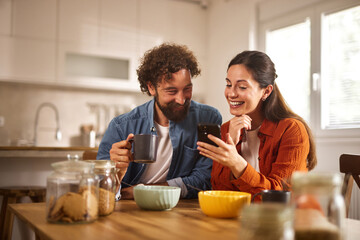 Two individuals smile and engage with each other while sitting at a wooden table in a bright home office. One holds a coffee mug, and the other looks at a phone.
