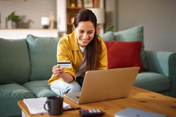 A woman sits on a sofa in a bright living room, holding a credit card and typing on her laptop. She...