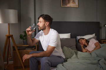 Man takes a sip from a glass of water while sitting on the edge of the bed, with his partner...
