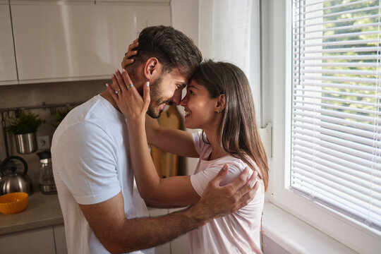 A couple embraces joyfully, foreheads touching, in a bright kitchen filled with morning light. They exude love and warmth, enjoying each other's presence.