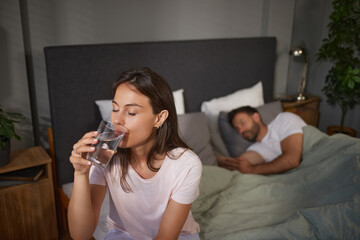 A woman sits on the edge of the bed, drinking a glass of water while her partner sleeps beside her in a dimly lit, comfortable bedroom.