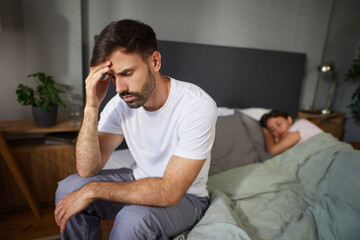 A man, visibly pained and thoughtful, sits on the edge of a bed while a woman lies turned away in...