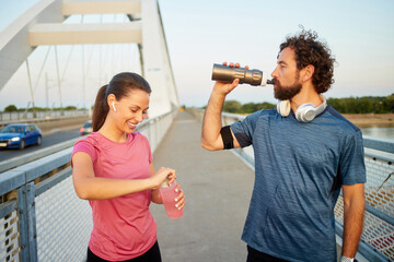 Jogging couple takes a break on a bridge during sunset. The woman opens a drink while the man...