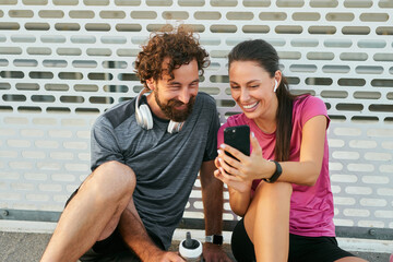 Two friends sit together on a track, smiling as they view their fitness app on a smartphone. They...