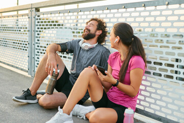 Two friends take a break after exercising, sitting together on a bridge. They share laughter while...