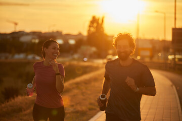 A man and woman are running side by side on a pathway during sunset, smiling and enjoying their...