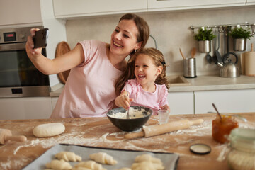 In a modern kitchen, a mother and her daughter smile brightly while taking a selfie amidst a baking...