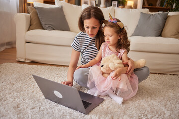 A mother and her daughter are sitting on the living room floor, engaging with a laptop together....