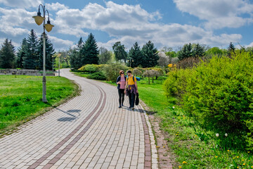 Couple walking on a paved path through a vibrant green park on a sunny spring day with trees,...
